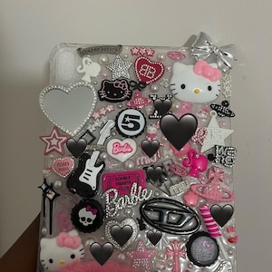 May include: A clear tablet case decorated with a collage of pink, black, and silver charms including Hello Kitty figures, Barbie logos, rhinestone hearts, stars, a guitar, a silver bow, and various brand emblems arranged across the entire surface.