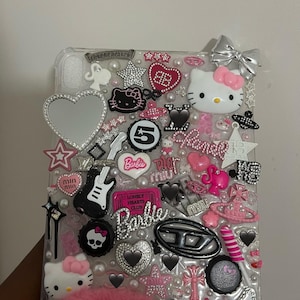 Könnte beinhalten: Transparente Handyhülle mit verschiedenen Charms, darunter Hello Kitty, Herzen, Sterne und das Wort "Barbie". Die Verzierungen sind in Pink, Schwarz, Silber und Weiß gehalten, mit einem flauschigen rosa Akzent unten.