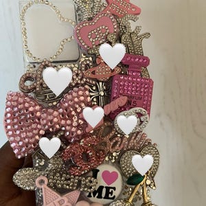 Può includere: Una custodia trasparente per telefono impreziosita da strass rosa e argento. La custodia presenta un grande fiocco rosa, un flacone di profumo, una stella e un design a forma di cuore. Sono visibili anche le parole "I LOVE ME".