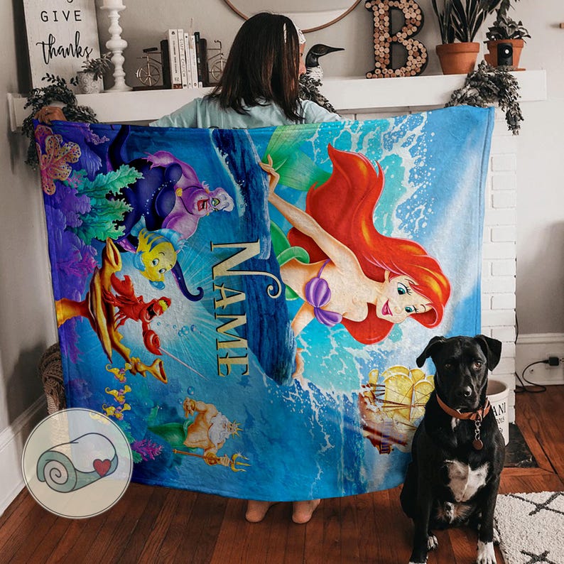 May include: A blue fleece blanket featuring a colorful illustration of Ariel from The Little Mermaid, with other characters and the word "NAME" printed on it. A black dog with a brown collar sits in front of the blanket.