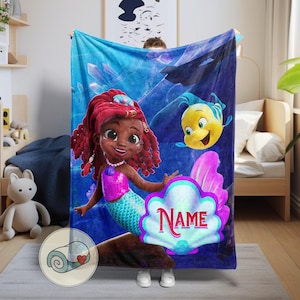 Peut inclure: Une couverture bleue avec une sir&egrave;ne de dessin anim&eacute; aux cheveux roux, un haut rose et une queue turquoise, accompagn&eacute;e d'un poisson jaune. La couverture a une zone en forme de coquillage avec le mot "NAME" en rose.