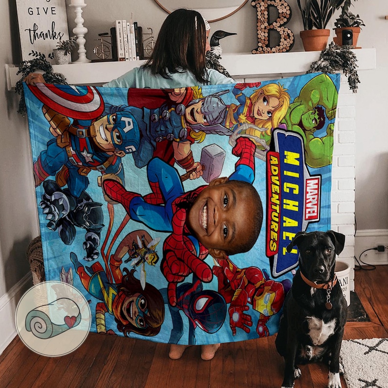 K&ouml;nnte beinhalten: Eine hellblaue Marvel Adventures-Decke mit verschiedenen Superhelden, darunter Captain America, Thor und Spider-Man. Die Decke hat ein personalisiertes Bild eines Kindes. Ein schwarzer Hund mit rotem Halsband sitzt vor der Decke.