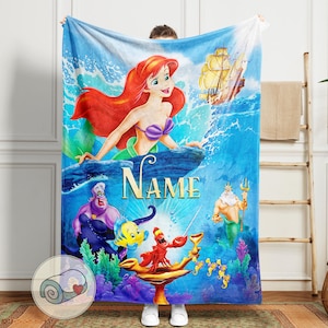 May include: A blue blanket featuring the Little Mermaid, Ariel, with red hair and a purple seashell bra, along with other characters from the movie. The word "NAME" is displayed in gold letters. A rolled-up blanket with a heart is in the corner.