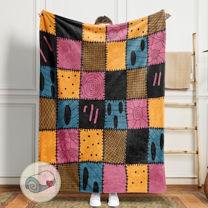 May include: A patchwork throw blanket featuring a variety of colorful squares in black, orange, pink, blue, and gold. Each square has a unique design, including spirals, dots, and stitched lines. The blanket is held up by a person wearing white shoes.