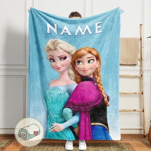 Puede incluir: Una manta azul claro con los personajes animados Elsa y Anna de la pel&iacute;cula Frozen. La palabra "NAME" est&aacute; impresa en blanco en la parte superior. La manta tiene una imagen enrollada de la manta con un coraz&oacute;n.