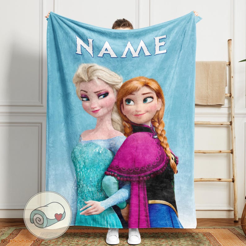 Puede incluir: Una manta azul claro con los personajes animados Elsa y Anna de la pel&iacute;cula Frozen. La palabra "NAME" est&aacute; impresa en blanco en la parte superior. La manta tiene una imagen enrollada de la manta con un coraz&oacute;n.