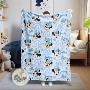 Puede incluir: Una manta azul claro con un estampado de Minnie Mouse, arco&iacute;ris, nubes, corazones y mariposas. El nombre "Jenny" est&aacute; escrito en todo el dise&ntilde;o. La manta est&aacute; siendo sostenida por una persona.