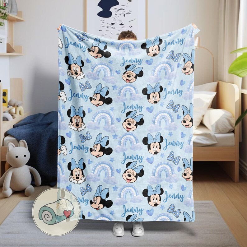 Puede incluir: Una manta azul claro con un estampado de Minnie Mouse, arco&iacute;ris, nubes, corazones y mariposas. El nombre "Jenny" est&aacute; escrito en todo el dise&ntilde;o. La manta est&aacute; siendo sostenida por una persona.