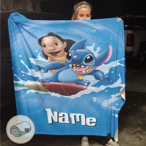 May include: A blue blanket featuring Stitch and Lilo surfing on a wave. The blanket has a blue background with white water splashes and the word "Name" in white block letters.