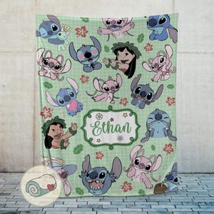 Puede incluir: Una manta verde claro con varios personajes de dibujos animados en azul y rosa, junto con detalles florales y de hojas. La manta tiene un fondo de patr&oacute;n de cuadr&iacute;cula y una etiqueta central con el nombre "Ethan".