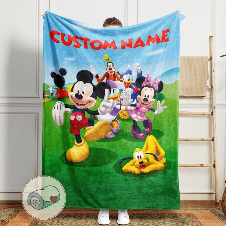 Pu&ograve; includere: Una coperta in pile blu con un'illustrazione colorata di Topolino, Minnie, Paperino, Pippo e Pluto. La coperta ha il testo "CUSTOM NAME" in lettere rosse. I personaggi sono ambientati su un campo verde e un cielo blu.