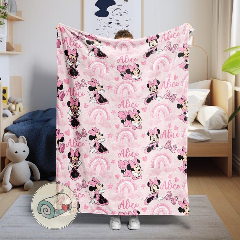 K&ouml;nnte beinhalten: Rosa Decke mit einem sich wiederholenden Muster von Minnie Mouse, Regenbogen, Sternen und dem Namen "Alice". Die Decke hat ein aufgerolltes Bild in der Ecke. Die Decke wird von einer Person gehalten.