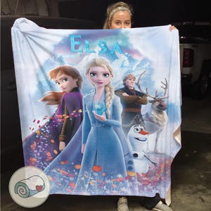 May include: A white blanket featuring characters from the animated film Frozen, including Elsa, Anna, Kristoff, Olaf, and Sven. The word "ELSA" is printed in blue at the top. The blanket has a colorful design with a winter theme.