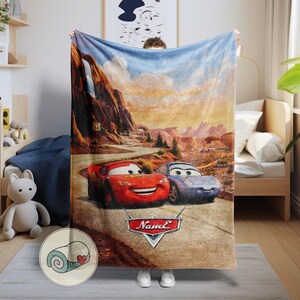 May include: A plush blanket featuring Lightning McQueen and Sally Carrera from the movie Cars, set against a desert landscape with Route 66 signage. The blanket is primarily blue, red, and brown.