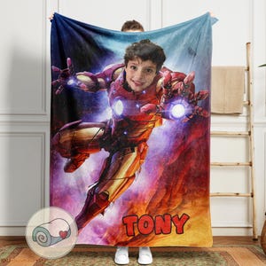 Peut inclure: Une couverture personnalis&eacute;e avec le visage d'un enfant superpos&eacute; &agrave; un design de costume d'Iron Man. La couverture est principalement rouge et or, avec le nom "TONY" en grandes lettres orange en bas. Le fond a un th&egrave;me cosmique.