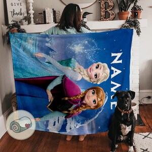 May include: A blue fleece blanket featuring Elsa and Anna from Frozen. The blanket has a personalized name section in white. The characters are depicted in their signature outfits against a snowy background. A rolled-up blanket with a heart logo is in the corner.