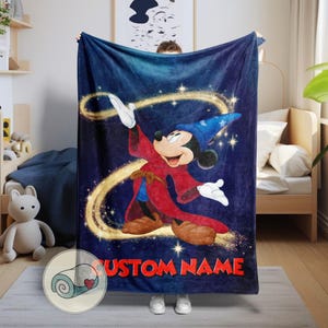 Op de afbeelding: Een marineblauwe deken met Mickey Mouse in een tovenaarsoutfit, met de tekst "CUSTOM NAME" in het rood. Het ontwerp bevat een blauwe tovenaarsmuts, een rode mantel en gouden steraccenten. De deken wordt door een persoon vastgehouden.