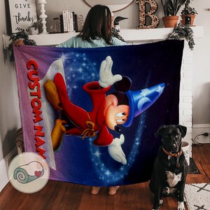 Pu&ograve; includere: Una grande coperta rettangolare con una vivace illustrazione di Topolino in abito da mago su uno sfondo blu stellato. La coperta ha una sezione viola e rosa con la scritta "CUSTOM NAME". Un cane nero &egrave; seduto davanti alla coperta.