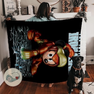 May include: Black blanket featuring a cartoon character in an Iron Man suit. The character's face is visible, with a red and gold suit design. The word "IRON MAN" is printed in blue. A black dog is sitting in front of the blanket.
