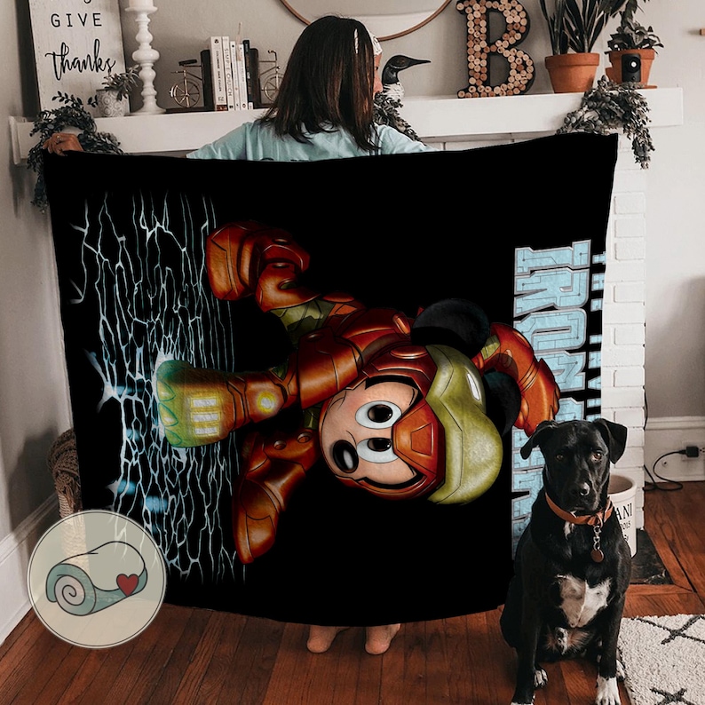 May include: Black blanket featuring a cartoon character in an Iron Man suit. The character's face is visible, with a red and gold suit design. The word "IRON MAN" is printed in blue. A black dog is sitting in front of the blanket.