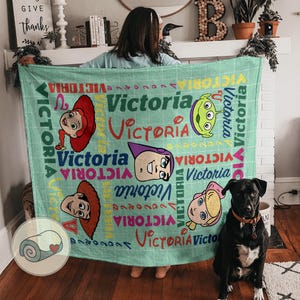 K&ouml;nnte beinhalten: Eine hellgr&uuml;ne Decke mit Illustrationen von Zeichentrickfiguren und dem Namen "Victoria" in verschiedenen Schriftarten und Farben. Die Decke hat ein Gittermuster und eine Grafik einer zusammengerollten Decke in der Ecke.