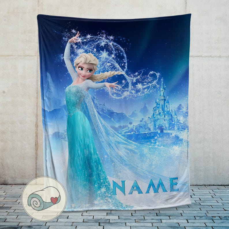Op de afbeelding: Een blauwe deken met een cartoonafbeelding van een vrouw in een vloeiende turquoise jurk met een kasteel op de achtergrond. Het woord "NAME" staat in het blauw onderaan. De deken wordt getoond tegen een grijze muur.