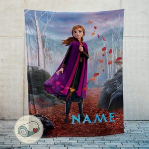Pode incluir: Um cobertor macio com uma personagem do filme Frozen, num cen&aacute;rio de floresta com folhas a cair. O cobertor tem uma capa roxa e rosa, e a palavra "NAME" &eacute; exibida em letras azuis.