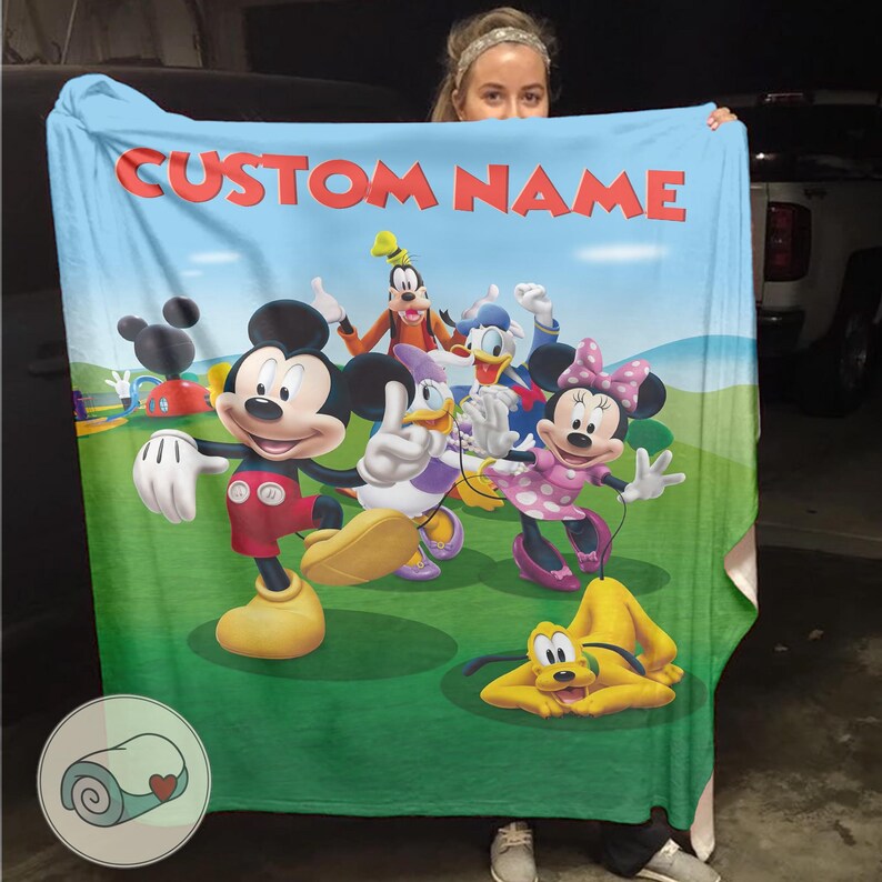Pu&ograve; includere: Una coperta azzurra con una vivace illustrazione di Topolino, Minnie, Paperino, Paperina, Pippo e Pluto. La coperta presenta la scritta "CUSTOM NAME" in rosso. Lo sfondo mostra un campo verde e un cielo blu.