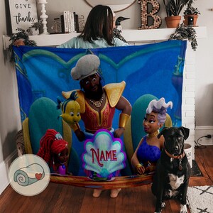 May include: A blue blanket with cartoon characters. The blanket depicts a man in a red and gold outfit, a yellow fish, a woman in a blue top, and a red-haired girl. A black dog sits in front of the blanket. The word "Name" is on the blanket.