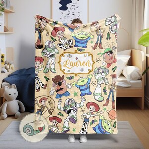 May include: A beige blanket featuring various characters from the Toy Story franchise. The blanket has a repeating pattern of Woody, Buzz Lightyear, Jessie, and the Little Green Men. The name "Lauren" is displayed in a decorative frame.