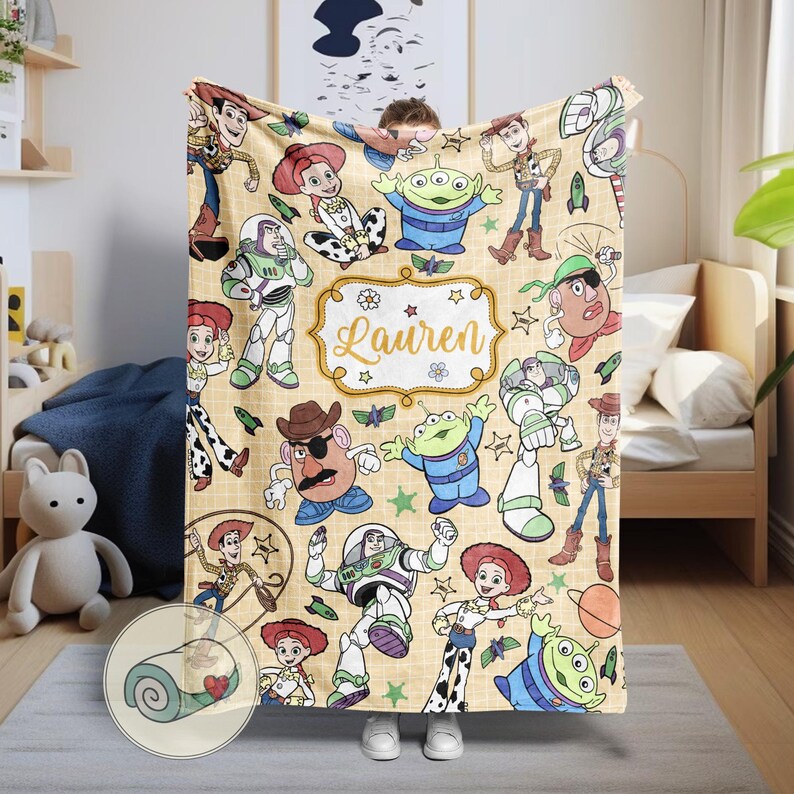 May include: A beige blanket featuring various characters from the Toy Story franchise. The blanket has a repeating pattern of Woody, Buzz Lightyear, Jessie, and the Little Green Men. The name "Lauren" is displayed in a decorative frame.