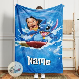 May include: Blue blanket featuring Stitch and Lilo surfing on a wave. The blanket has a blue background with the word "Name" in white. The image shows Stitch with a big smile and Lilo smiling. The blanket is soft and cozy.