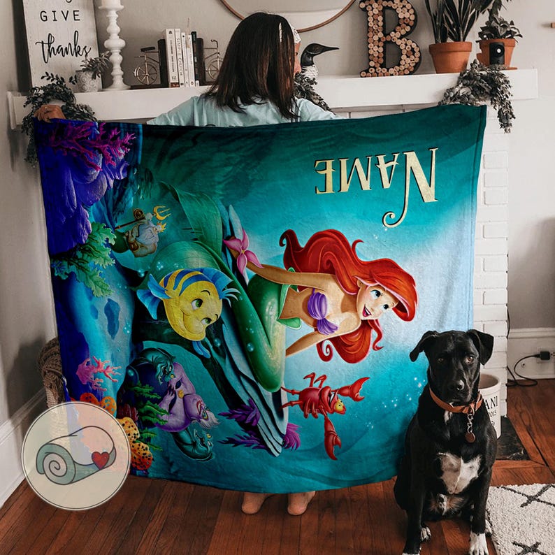 Puede incluir: Una manta turquesa con una sirena de pelo rojo, top morado y cola verde, junto a Flounder y Sebasti&aacute;n. La manta tiene la palabra "NAME" en letras doradas y est&aacute; rodeada de una escena submarina.