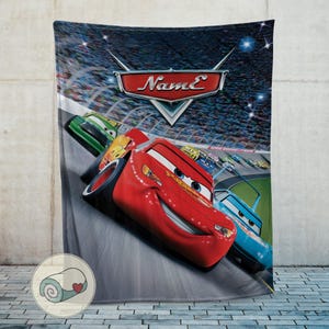 May include: A plush blanket featuring a vibrant image of the animated movie "Cars." The design showcases the character Lightning McQueen racing on a track, with the movie's logo at the top. The blanket is primarily red, blue, and green.