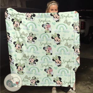 K&ouml;nnte beinhalten: Eine hellgr&uuml;ne Decke mit einem Muster aus Minnie-Maus-Illustrationen, Schleifen, Regenbogen und dem Namen "Sophia" in Schreibschrift. Die Decke hat ein weiches, pastellfarbenes Farbschema mit wei&szlig;en Wolken und gr&uuml;nen Akzenten.