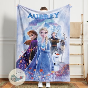 May include: A plush blanket featuring characters from the movie Frozen, including Elsa, Anna, Kristoff, Olaf, and Sven. The blanket is light blue with the name "AUBREY" in large, blue letters at the top. The blanket is held up by a person.