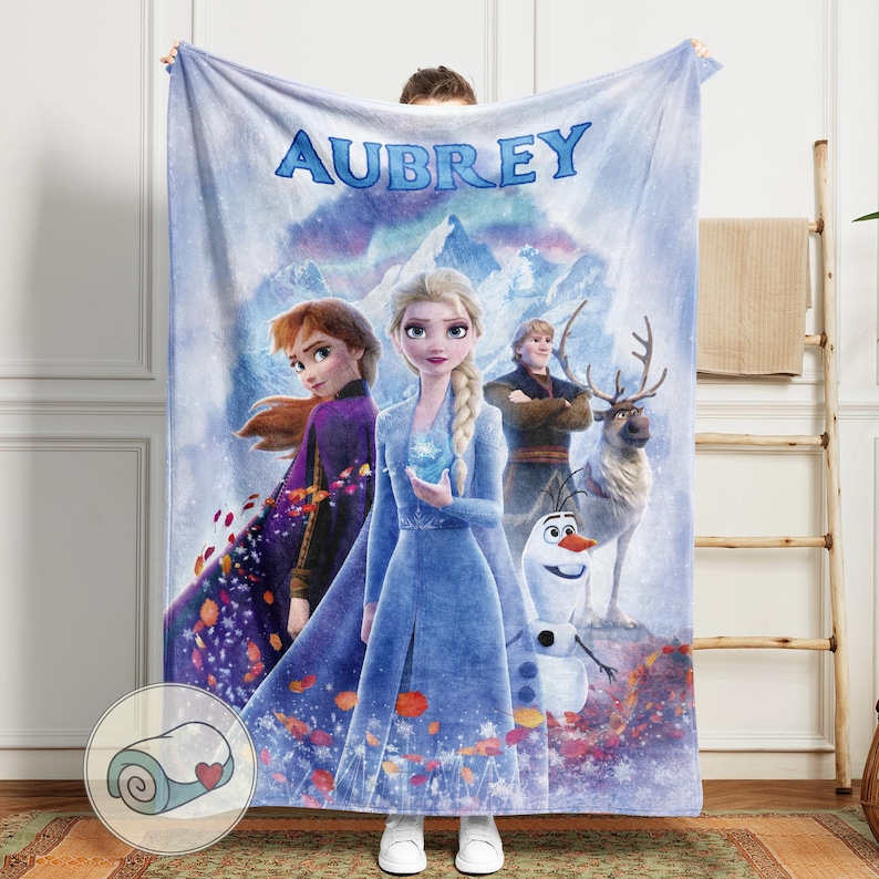 May include: A plush blanket featuring characters from the movie Frozen, including Elsa, Anna, Kristoff, Olaf, and Sven. The blanket is light blue with the name "AUBREY" in large, blue letters at the top. The blanket is held up by a person.