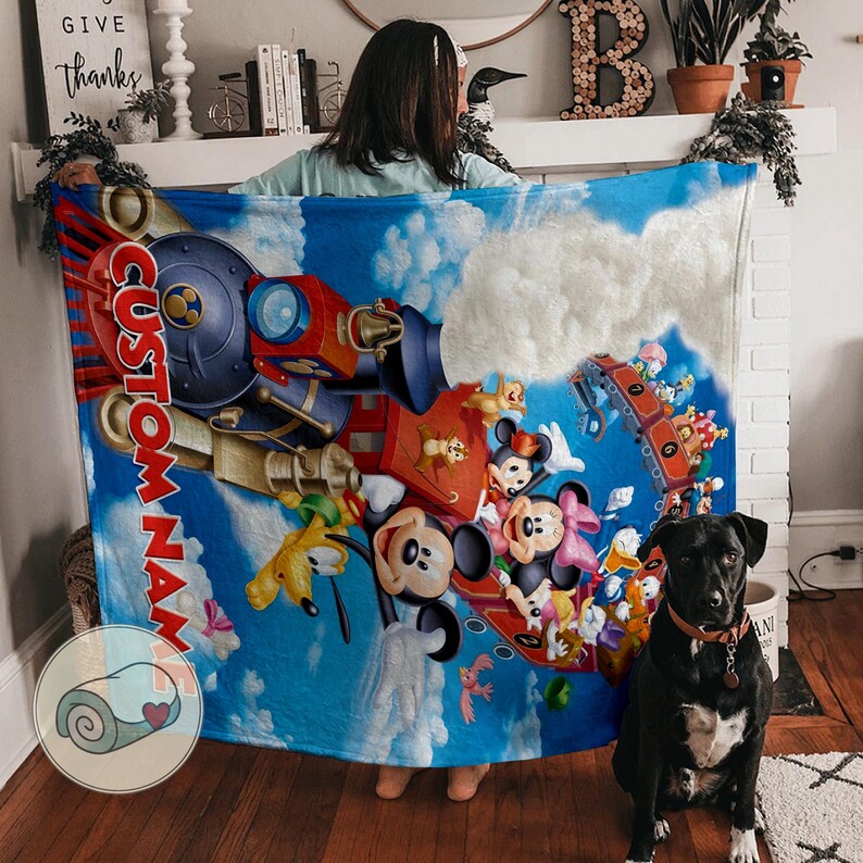 May include: A blue blanket featuring a train design with Mickey Mouse, Minnie Mouse, and other Disney characters. The blanket has the text "CUSTOM NAME" in red. A black dog sits in front of the blanket.