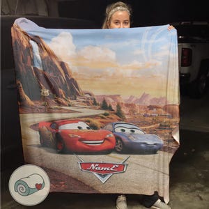 May include: A large, rectangular blanket featuring Lightning McQueen and Sally Carrera from the movie Cars. The blanket showcases a scenic desert landscape with Route 66 signage and a waterfall. The Cars logo is visible.