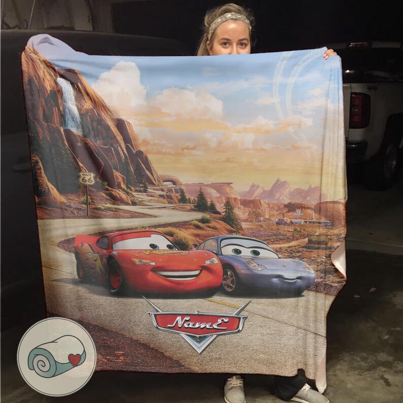 May include: A large, rectangular blanket featuring Lightning McQueen and Sally Carrera from the movie Cars. The blanket showcases a scenic desert landscape with Route 66 signage and a waterfall. The Cars logo is visible.
