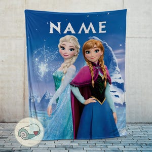 May include: A blue blanket featuring the characters Elsa and Anna from the movie Frozen, with the word "NAME" in white at the top. The blanket has a wintery background with snowflakes and mountains.