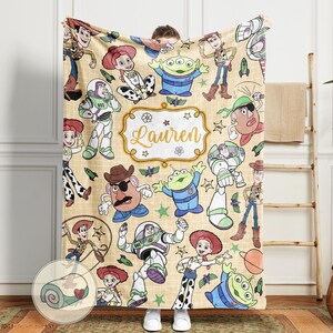 May include: A beige blanket featuring various Toy Story characters, including Woody, Buzz Lightyear, and Jessie. The blanket has a repeating pattern of the characters and includes the name "Lauren" in a decorative frame.