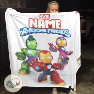 Op de afbeelding: Witte Marvel-deken met de tekst "NAME and Awesome Friends" en cartoon illustraties van Iron Man, Hulk en een ander personage. De deken heeft een opgerold beeld in de linker benedenhoek.