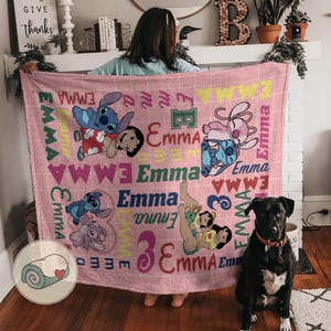 May include: A pink blanket featuring cartoon characters and the name "Emma" in various fonts. The blanket has a grid pattern and includes images of Stitch and Lilo. A black dog with a collar sits nearby.
