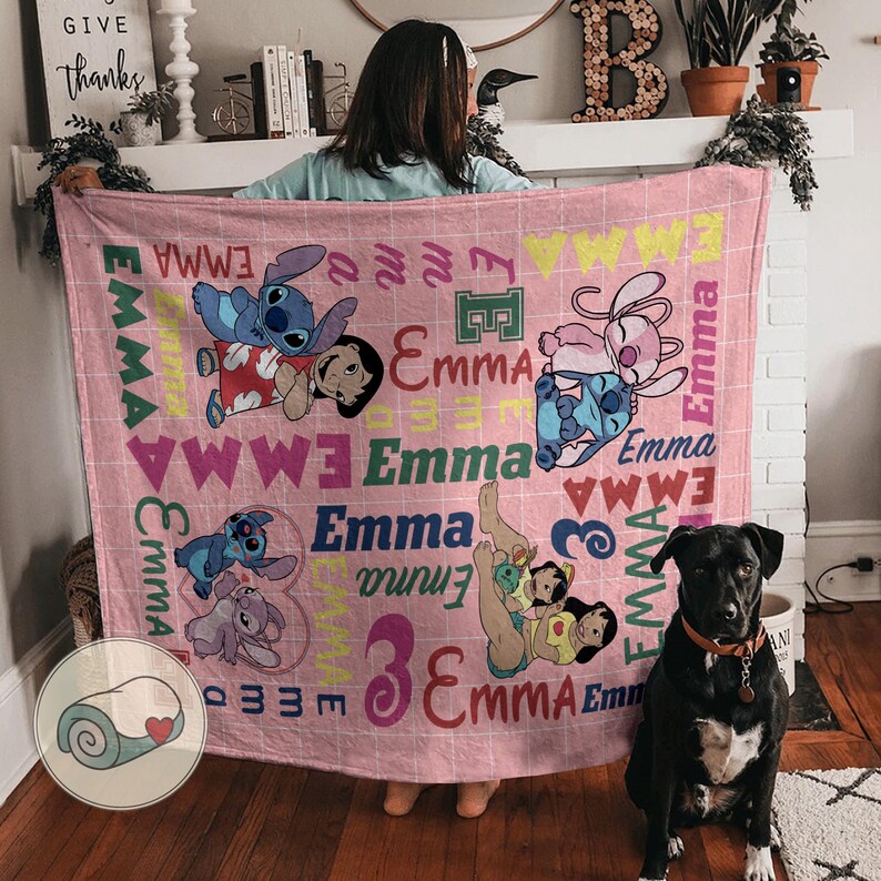 May include: A pink blanket featuring cartoon characters and the name "Emma" in various fonts. The blanket has a grid pattern and includes images of Stitch and Lilo. A black dog with a collar sits nearby.