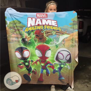 Pode incluir: Uma manta Marvel "Name and His Amazing Friends". A manta apresenta uma ilustra&ccedil;&atilde;o colorida do Homem-Aranha, Miles Morales e Spider-Gwen, juntamente com outros personagens. A manta tem um fundo azul claro com uma paisagem de desenho animado.