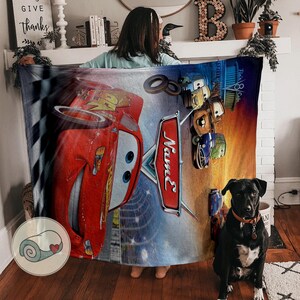 May include: A large, rectangular blanket featuring Lightning McQueen from the Cars movie. The blanket has a vibrant design with a red race car, the word "Name" in a stylized font, and other characters from the movie. The background includes a checkered flag and a sunset.