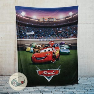 May include: A plush blanket featuring characters from the animated movie Cars. The blanket showcases Lightning McQueen and other race cars on a green field in front of a stadium filled with spectators. The blanket has a dark blue sky background.