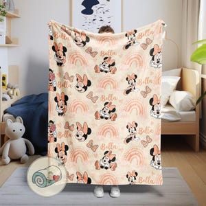 May include: A beige blanket featuring a repeating pattern of Minnie Mouse, bows, rainbows, and the name "Bella" in a soft, muted color palette. The blanket is held up by a person wearing white shoes.