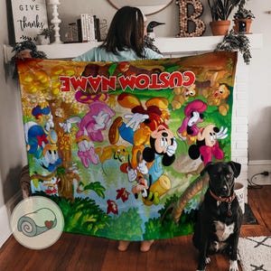May include: A colorful blanket featuring cartoon characters, including Mickey Mouse, Donald Duck, and Goofy, with the text "CUSTOM NAME" in red. The blanket has a green and yellow background with a forest scene.